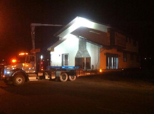 A McConnell Building Mover semi hauling a house on dollies in the night is illuminated by flood lamps.